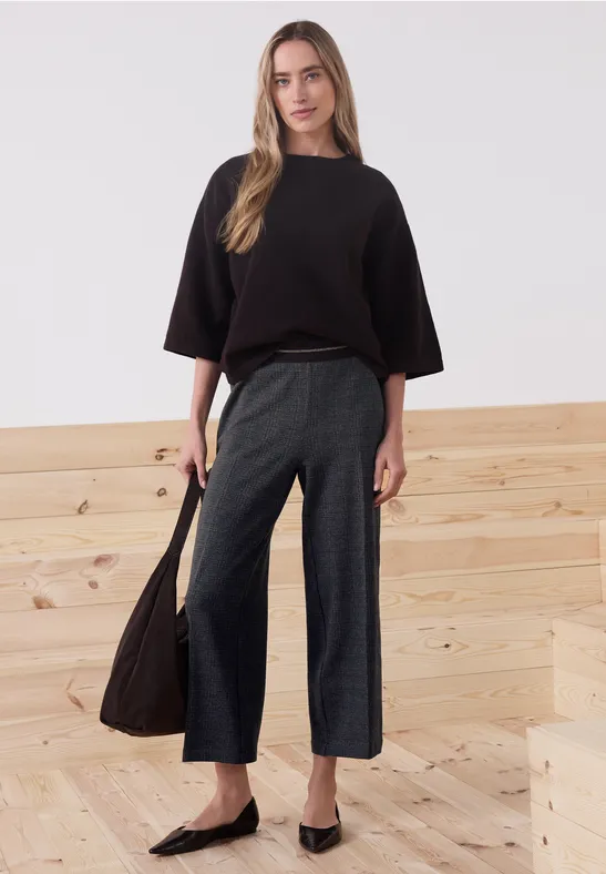 Frau in schwarzem Oversize-Pullover, weiter grauer Hose und schwarzen Ballerinas, vor hellem Holztreppen-Hintergrund.