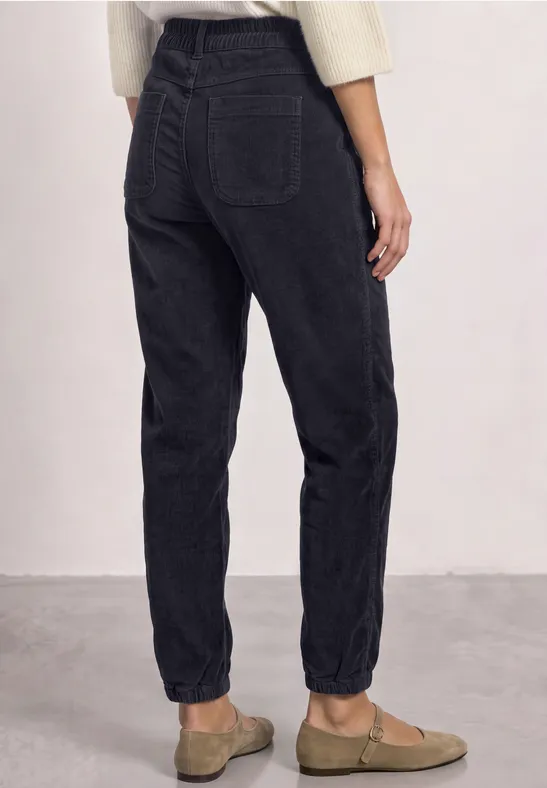 Rückansicht einer Person in schwarzen, hoch taillierten, lässigen Jeans mit aufgerolltem Saum und beige Slippern.