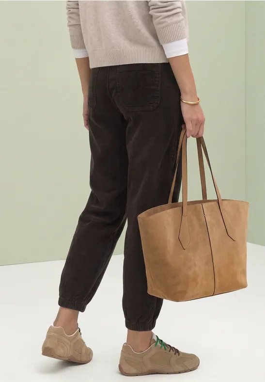 Vrouw met beige trui, bruine corduroy broek, lichte sneakers en grote suède shopper.