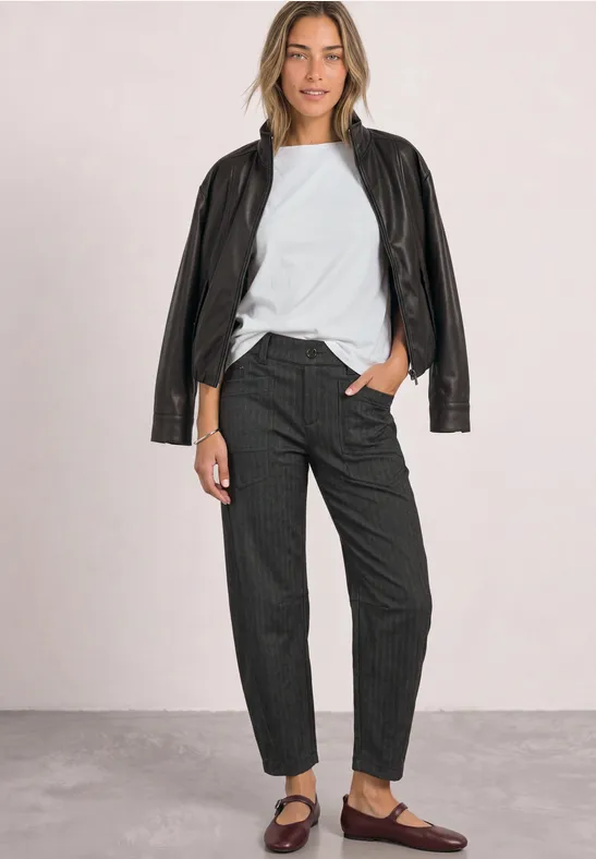 Frau in weißem Shirt, schwarzer Lederjacke, dunklen Hosen und braunen Loafers vor hellem Hintergrund.