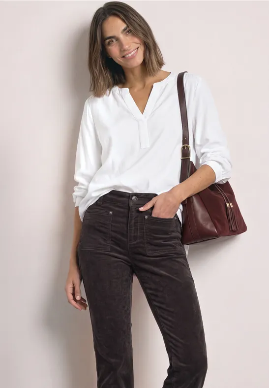Frau in weißer Bluse und brauner Cordhose mit Schultertasche, lächelnd vor hellem Hintergrund.