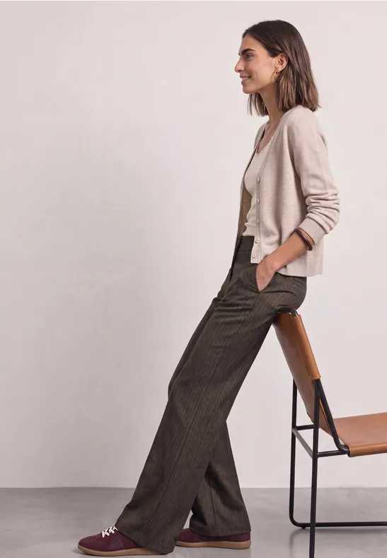 Eine Frau sitzt seitlich auf einem Stuhl, trägt einen hellen Cardigan, weißes Top und weite braune Bundfaltenhose.