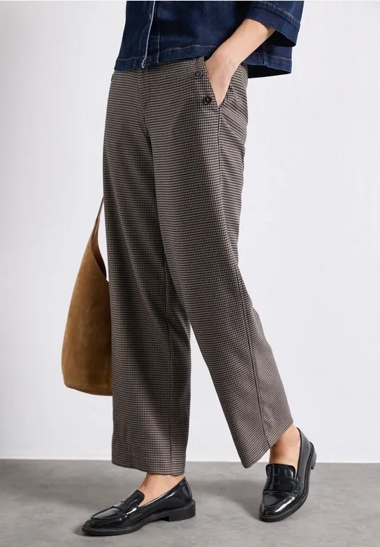 Person trägt graue Stoffhose, schwarze Loafers und hält eine braune Tasche.