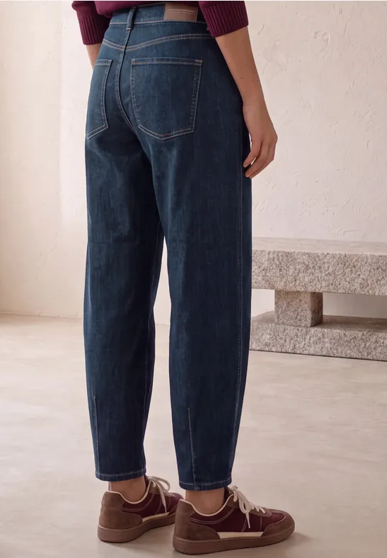 Une personne vue de dos porte un jean taille haute bleu foncé et des baskets marron sur fond clair.