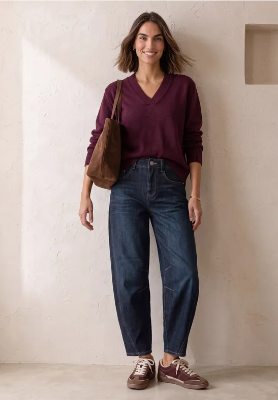 Une femme souriante porte un pull bordeaux, un jean bleu et un sac marron contre un mur.