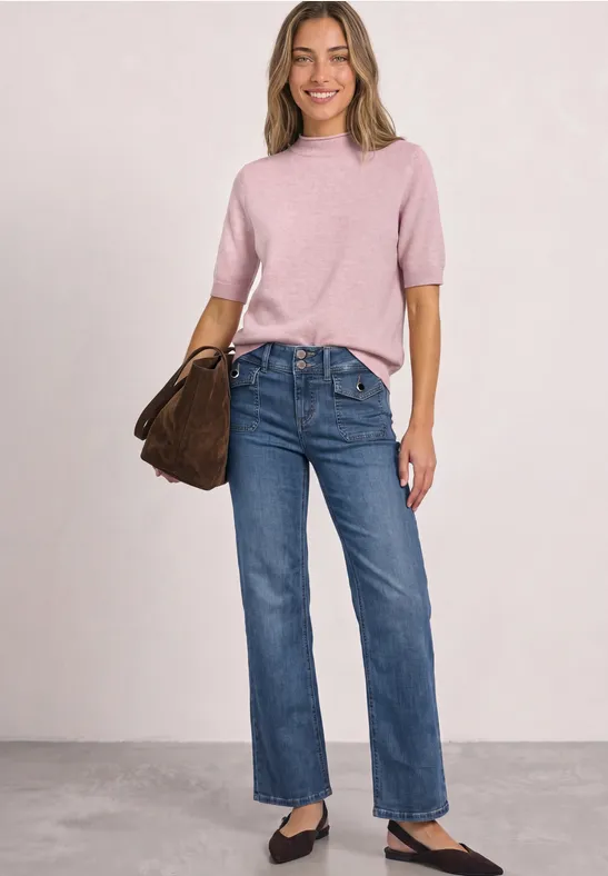 Frau trägt rosa Kurzarm-Pullover, blaue Jeans, schwarze Ballerinas und braune Tasche.