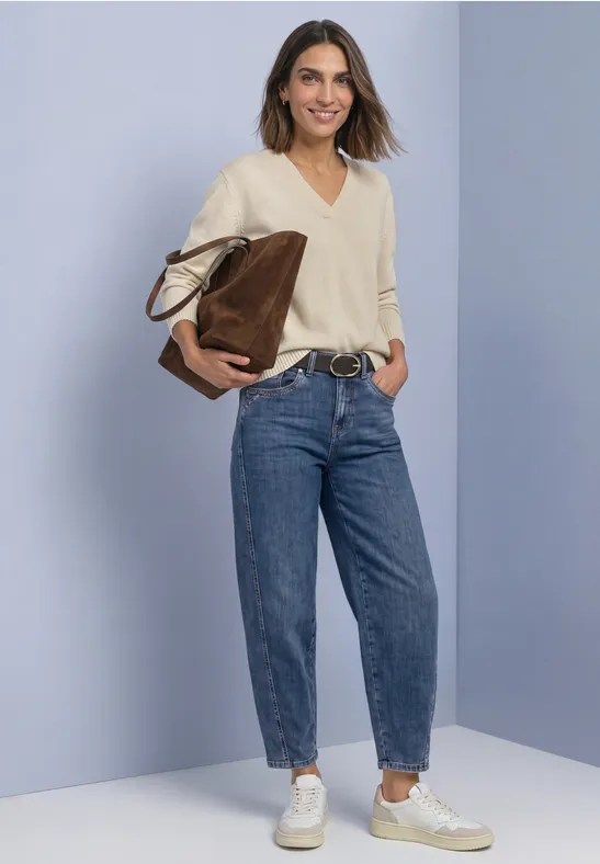 Frau im hellen Pullover und Jeans mit Sneakern und großer Tasche vor einfarbigem Hintergrund.
