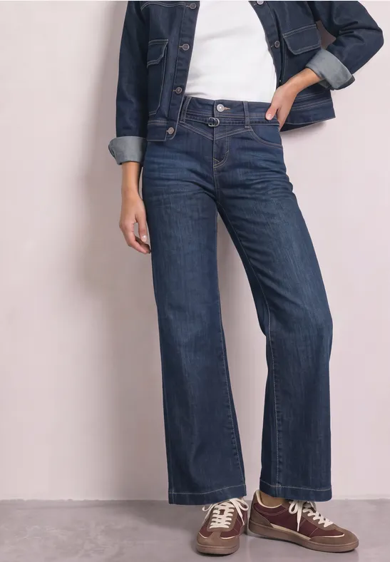 Modell trägt hoch sitzende, dunkelblaue Schlagjeans, weißes T-Shirt, Jeansjacke und braune Sneaker.