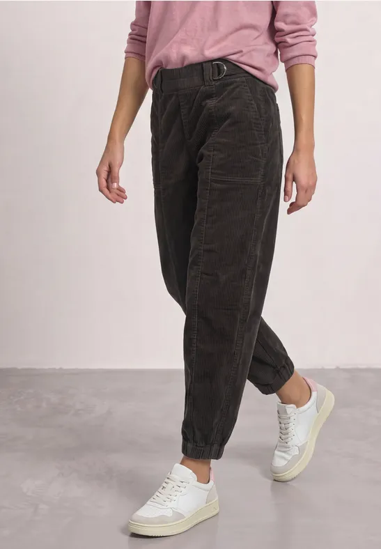 Donkerbruine ribfluwelen broek met losse pasvorm opgerold bij de enkels, gecombineerd met een roze trui en witte sneakers.