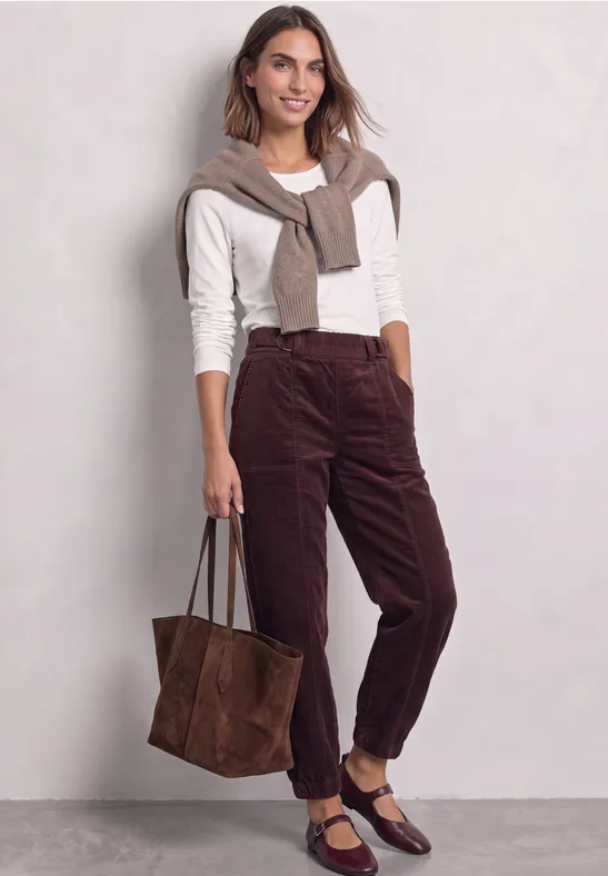 Frau in weißem Pullover, brauner Cordhose und passenden Accessoires mit großer Wildledertasche.