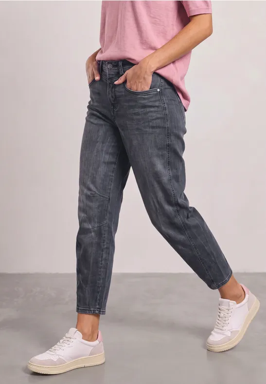 Person in taillierten dunkelgrauen Jeans, weißen Sneakern und rosa Shirt vor neutralem Hintergrund.