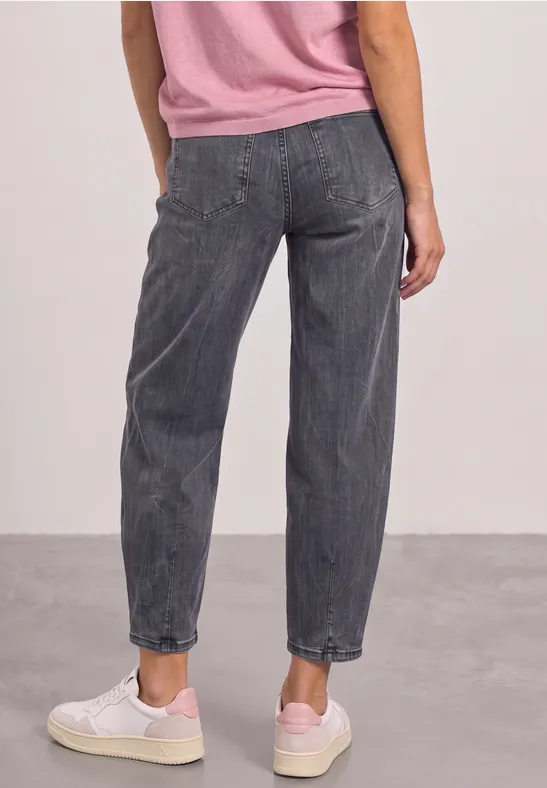 Rückansicht einer Person in lockeren grauen Jeans, rosa Oberteil und weißen Sneakers vor neutralem Hintergrund.