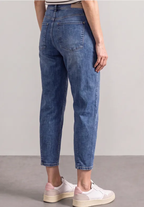 Rückenansicht einer Person in hochtaillierten blauen Jeans und weißen Sneakern vor neutralem Hintergrund.