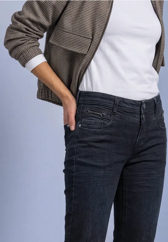 Eine Person in weißem T-Shirt, olivgrüner Jacke und dunklen Jeans steckt die Hand in die Hosentasche.