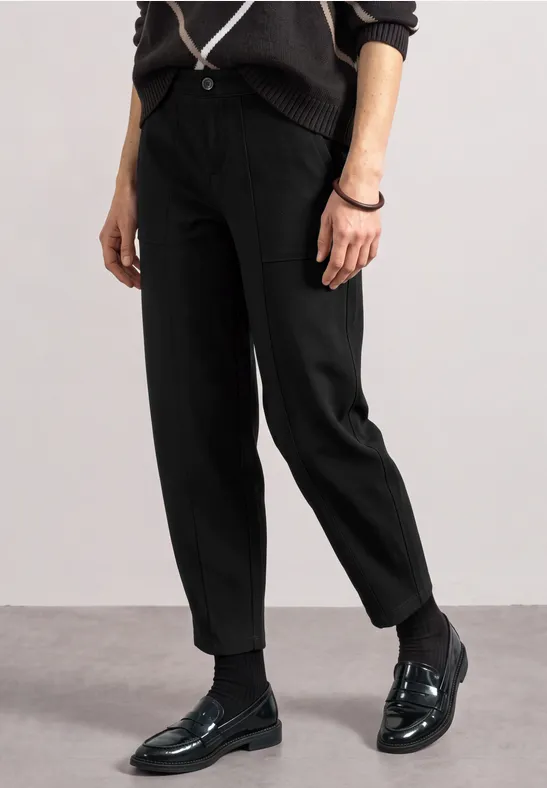 Eine Person trägt schwarze Stoffhose und glänzende schwarze Loafers auf grauem Boden.