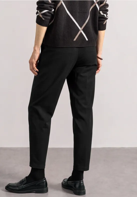 Person von hinten in schwarzen Hosen, schwarzen Lederschuhen und dunklem Pullover mit X-Muster.