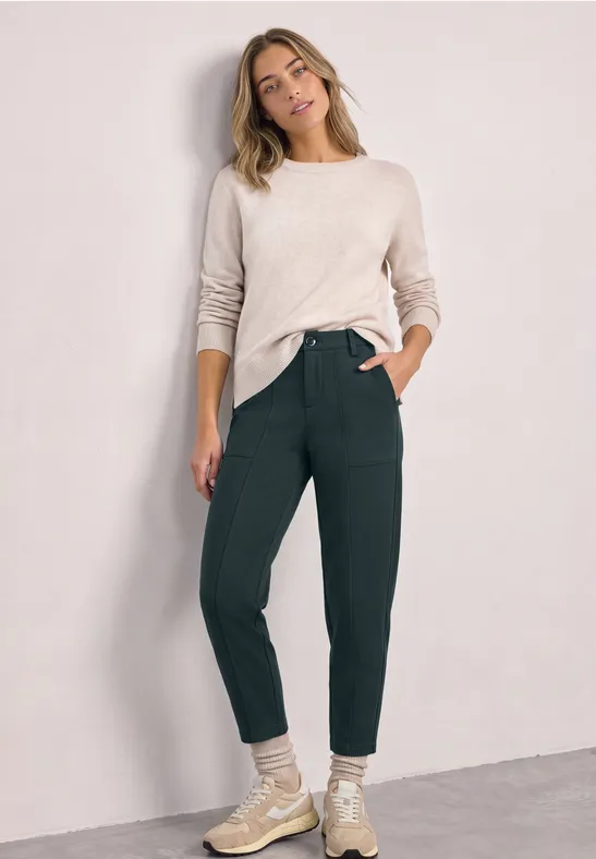 Eine Frau steht vor einer hellen Wand, trägt beigen Pullover, grüne Hose und helle Schuhe, lächelnd.