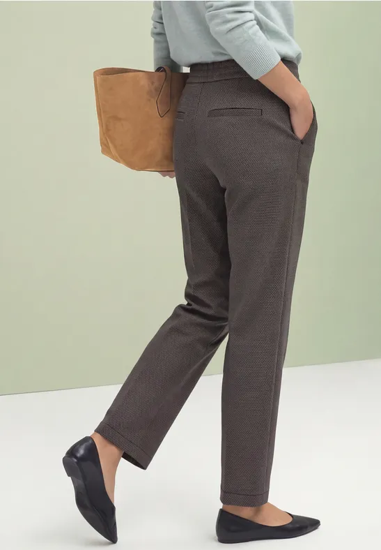Frau trägt graubraune, gerade geschnittene Hose, schwarze Ballerinas, hält braune Tasche.