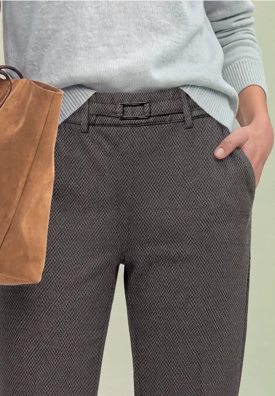 Nahaufnahme: graue, strukturierte Hose mit Gürtel, Hand in Tasche, hellblauer Pullover, braune Wildledertasche.