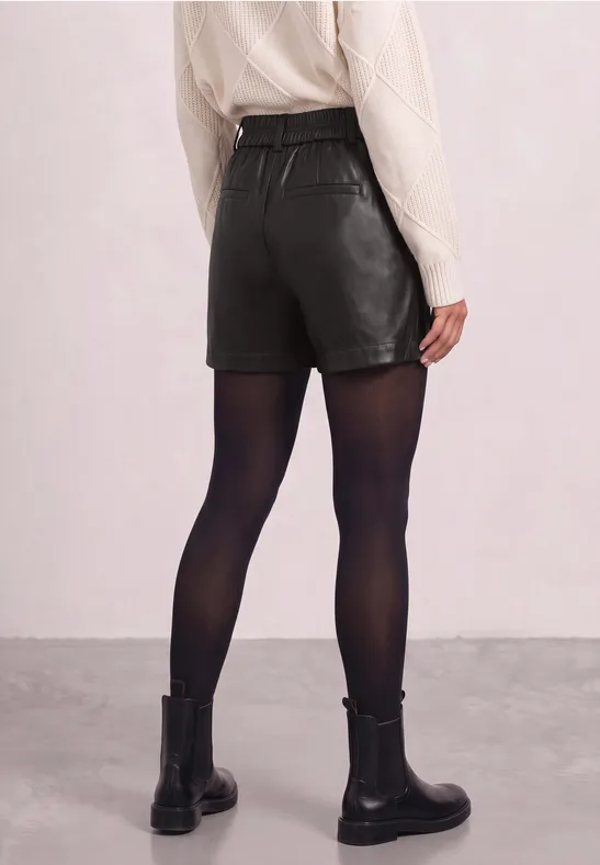 Rückansicht einer Person mit beigem Strickpullover, schwarzen Leder-Shorts, schwarzen Strumpfhosen und schwarzen Boots.