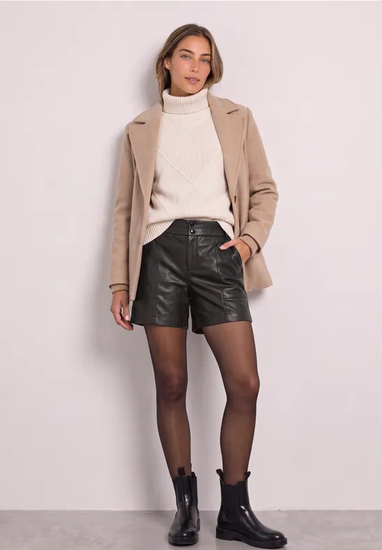 Frau in beigem Mantel, weißem Rollkragenpullover, schwarzen Leder-Shorts, Strumpfhose und schwarzen Stiefeletten