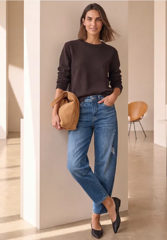 Frau in braunem Pullover und Jeans mit Strohhut in heller, minimalistischer Umgebung.