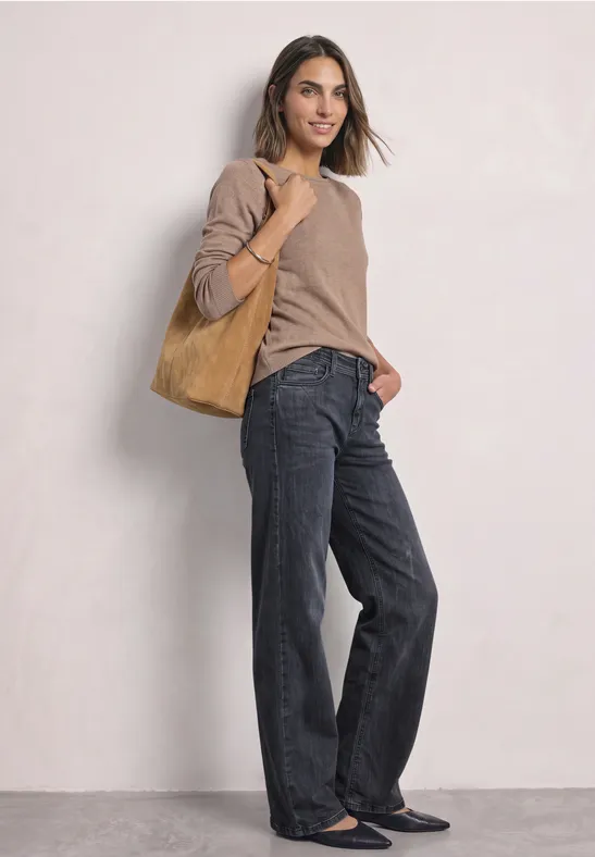 Femme en pull beige, jean large noir et ballerines, portant un cabas en daim marron devant un fond clair.