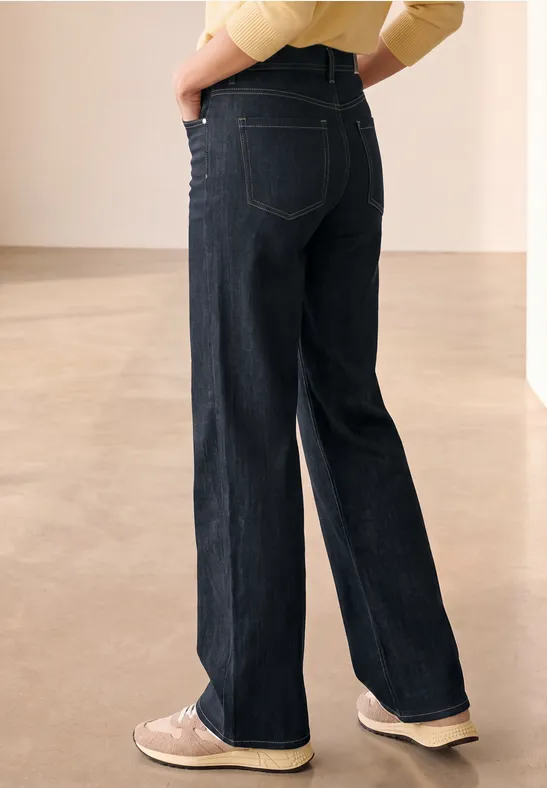 Rückansicht einer Person in dunklen, weiten Jeans und hellen Sneakers vor neutralem Hintergrund.