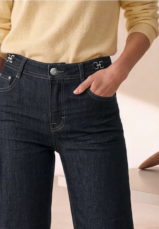 Person trägt dunkle High-Waist-Jeans und hellbeigen Pullover, Hand in Tasche, minimalistische Wohnraum-Kulisse.