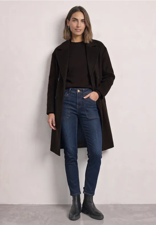 Frau trägt schwarzen Mantel, dunklen Pullover, blaue Jeans und schwarze Stiefeletten vor hellem Hintergrund.