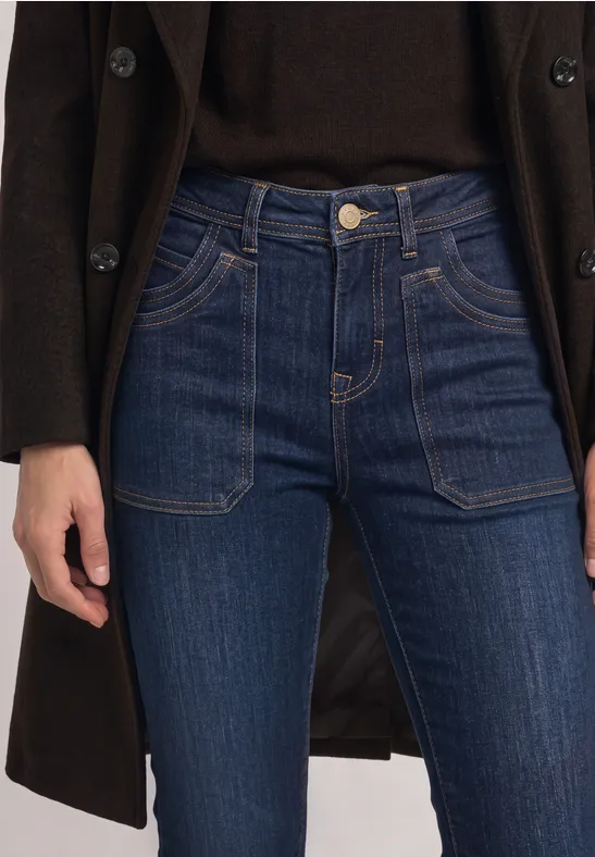 Frau in dunkelblauer Jeans mit aufgesetzten Taschen, dunklem Oberteil und langem Mantel.