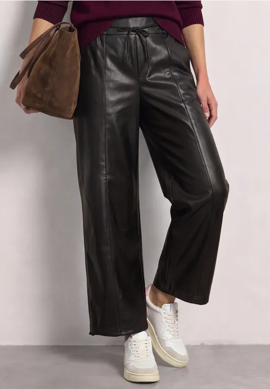 Style tendance : pantalon en cuir noir ample, baskets blanches et sac en daim marron sur fond neutre.