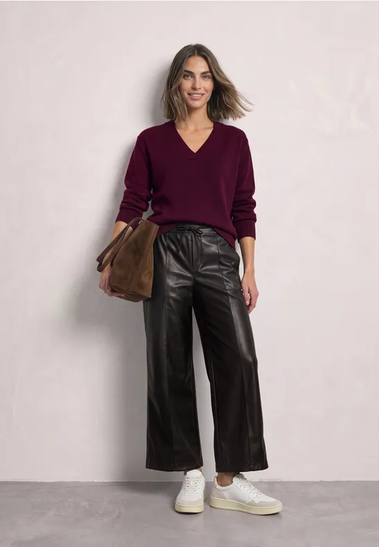 Femme devant fond clair, pantalon large en cuir noir, pull bordeaux à col V, baskets blanches et sac marron.