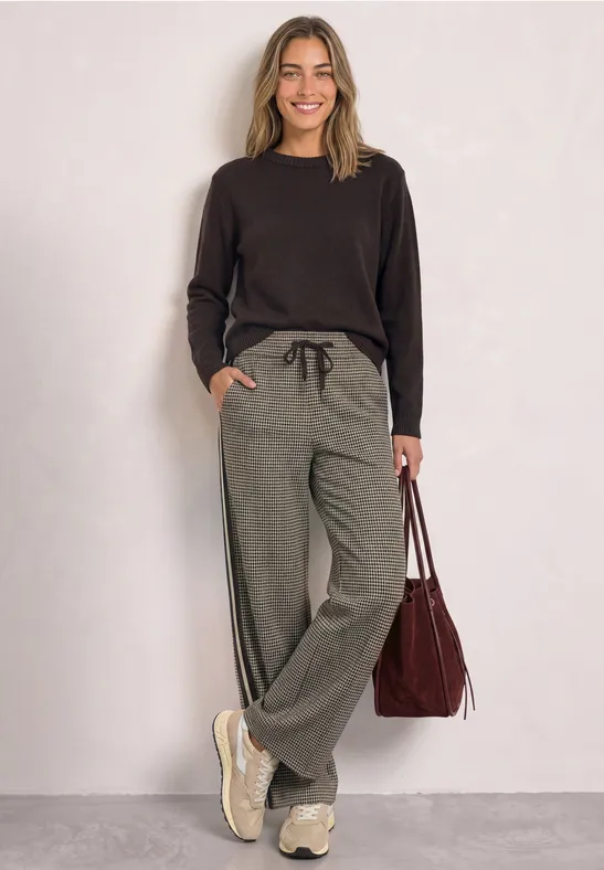 Lächelnde Frau in schwarzem Pullover, grauen Jogginghosen und Sneakers hält rote Tasche in lockerer Pose.
