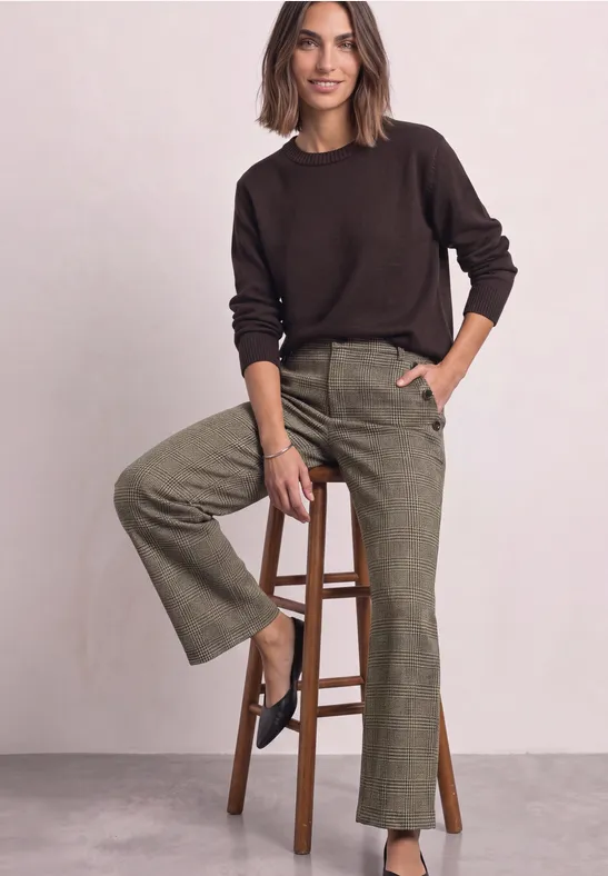 Eine Frau sitzt auf einem Hocker, trägt braunen Pullover und grüne Cordhose vor neutralem Hintergrund.