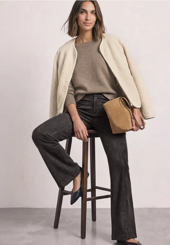 Frau im lässigen Outfit: helle Jacke über den Schultern, brauner Pullover, dunkle Jeans, braune Tasche auf dem Schoß.