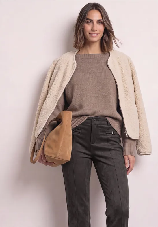Frau in beigem Teddy-Mantel, braunem Pullover und dunkelbrauner Hose mit brauner Tasche vor hellem Hintergrund.