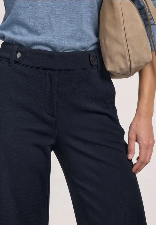 Close-up einer Taille mit dunkelblauer Hose, hellblauem Oberteil und beiger Handtasche.
