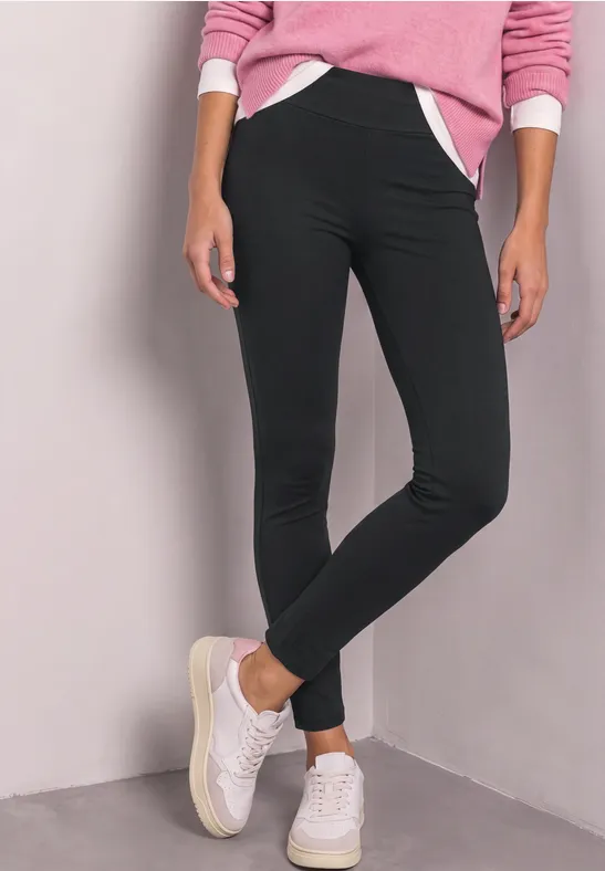 Frau in schwarzer Leggings, rosa Pullover und weißen Sneakern steht vor hellgrauer Wand.