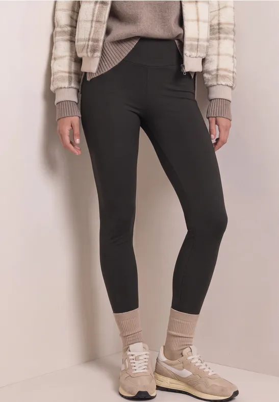 Schlanke Person in schwarzen Leggings, beigen Socken, Sneakern und karierter Jacke vor hellem Hintergrund.