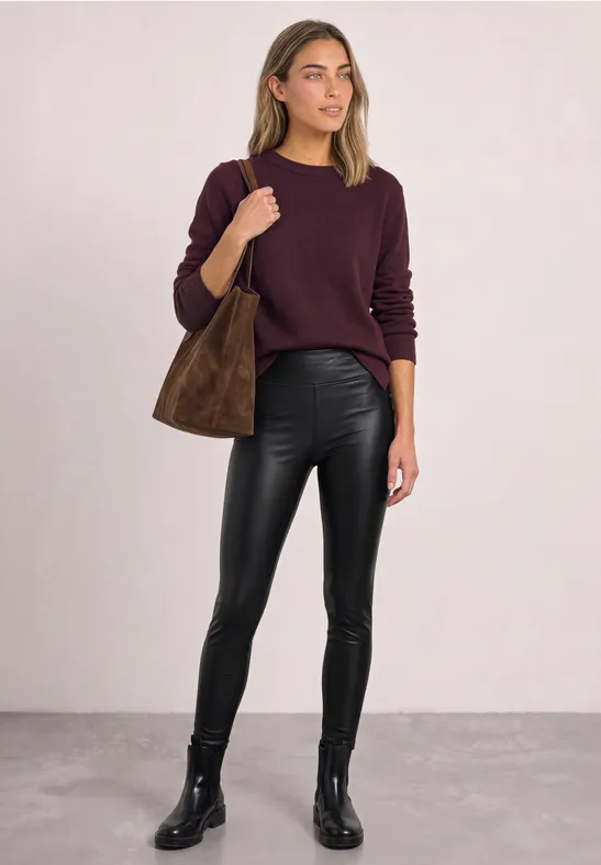 Eine Frau steht in schwarzer Lederhose, schwarzen Stiefeln, bordeauxrotem Pullover und brauner Tasche.