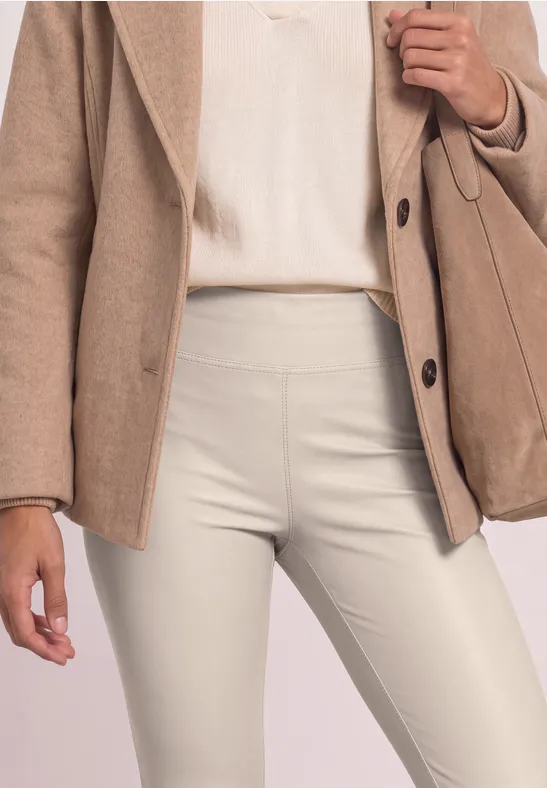 Close-up van een persoon met een beige blazer, licht topje en crèmekleurige, nauwsluitende broek met bijpassende tas.