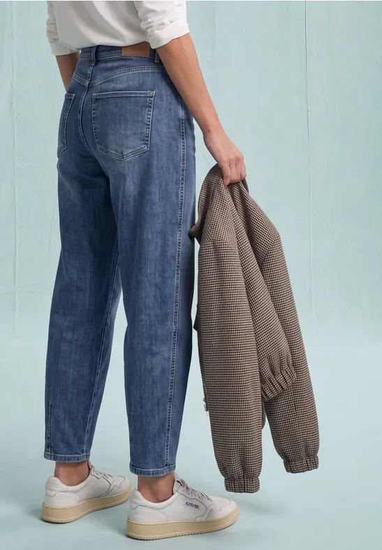 Person von hinten in lockeren Jeans und weißen Sneakern, hält eine graubraune Jacke in der Hand.
