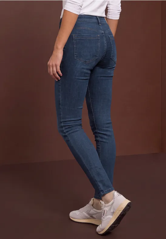Person trägt dunkelblaue Skinny-Jeans und weiße Sneaker vor braunem Hintergrund.