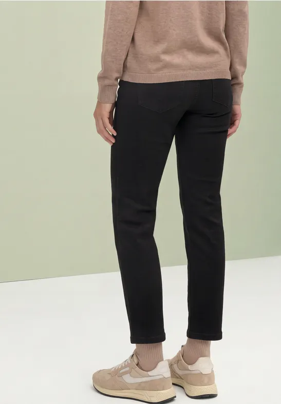 Person von hinten trägt schwarze Hose, beige Sneaker und braunen Pullover vor grün-weißem Hintergrund.
