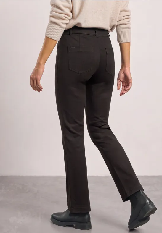 Persoon van achteren in een donkere broek, beige trui en zwarte laarzen tegen een lichte achtergrond.