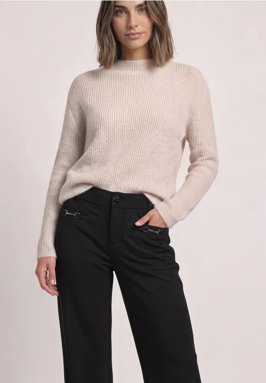Frau in beigem Pullover und schwarzer Hose mit Reißverschluss-Detail, eleganter Casual-Look.
