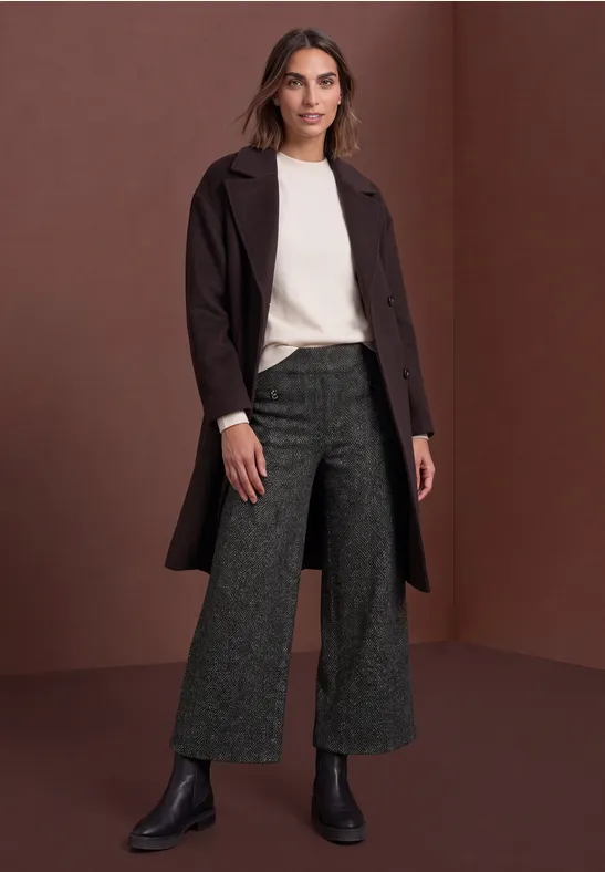 Frau in cremefarbenem Pullover, dunkelbraunem Mantel, weiten grauen Hosen und schwarzen Boots vor braunem Hintergrund.
