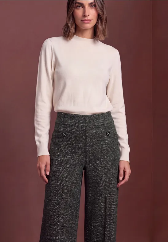 Frau in cremefarbenem Langarmshirt und eleganter, weiter, dunkelgrüner Tweed-Hose vor schlichtem Hintergrund.