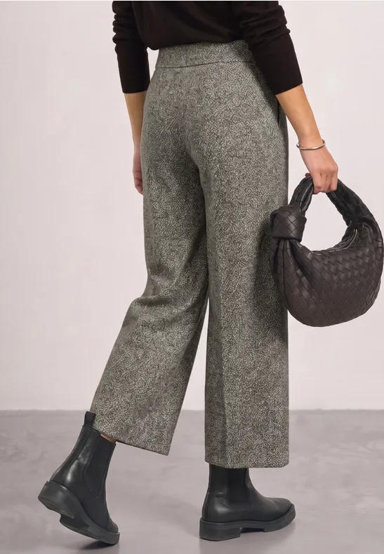 Frau trägt graue, hochgeschnittene Hose, schwarzen Pullover und Boots, hält eine schwarze geflochtene Tasche.